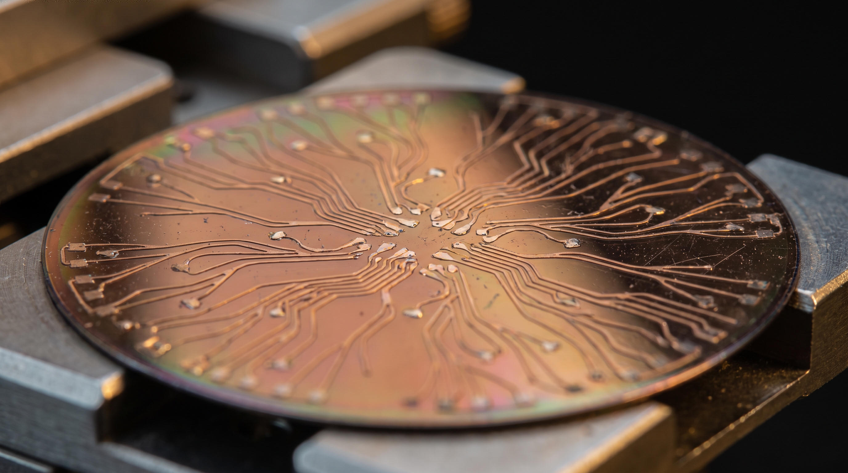 A copper synthetic neural substrate on a probe station: branching conductive traces radiate from a central hub across an iridescent wafer, resembling a nervous system yet clearly fabricated.